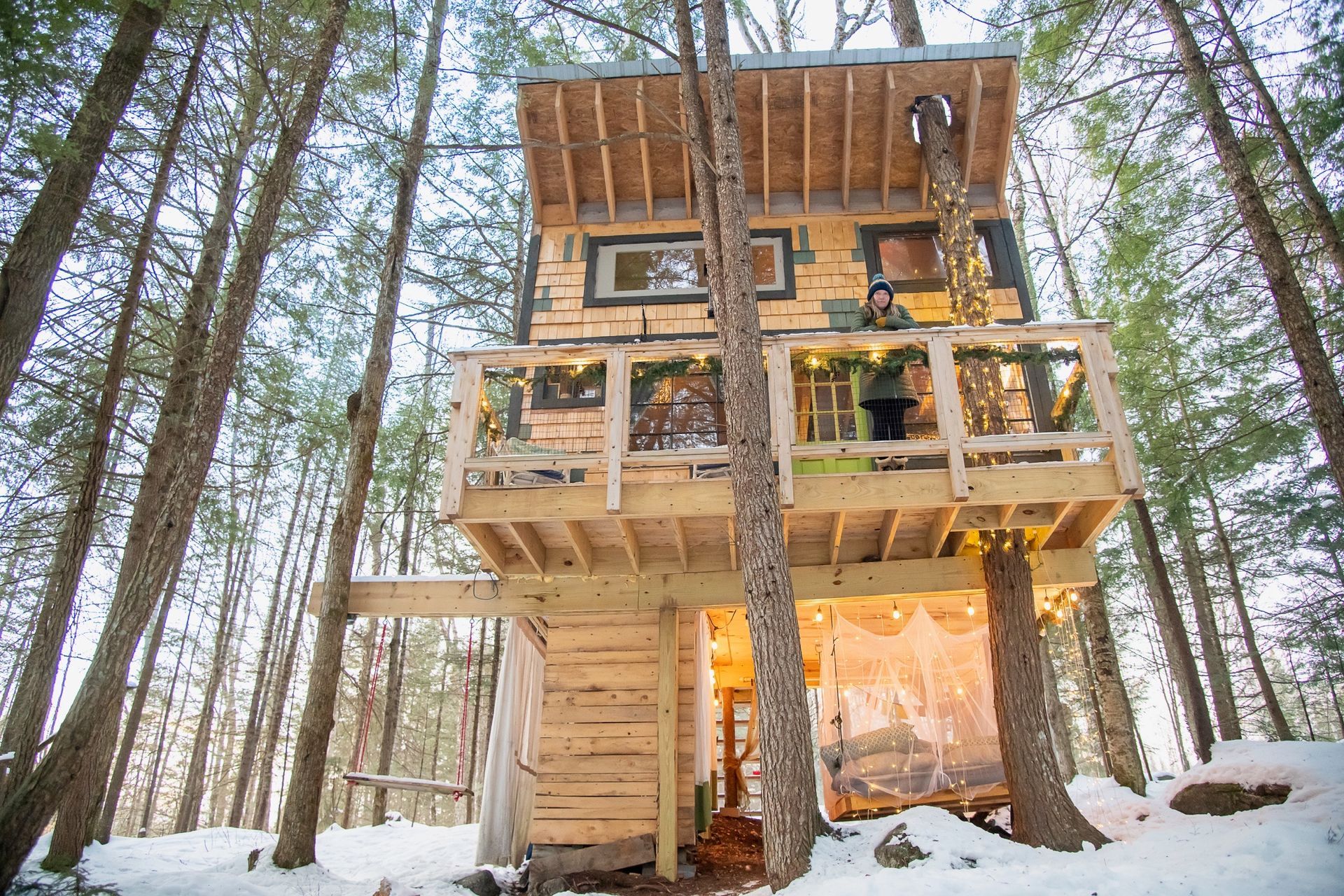 A multi-story treehouse in a snowy forest; person on balcony with string lights.