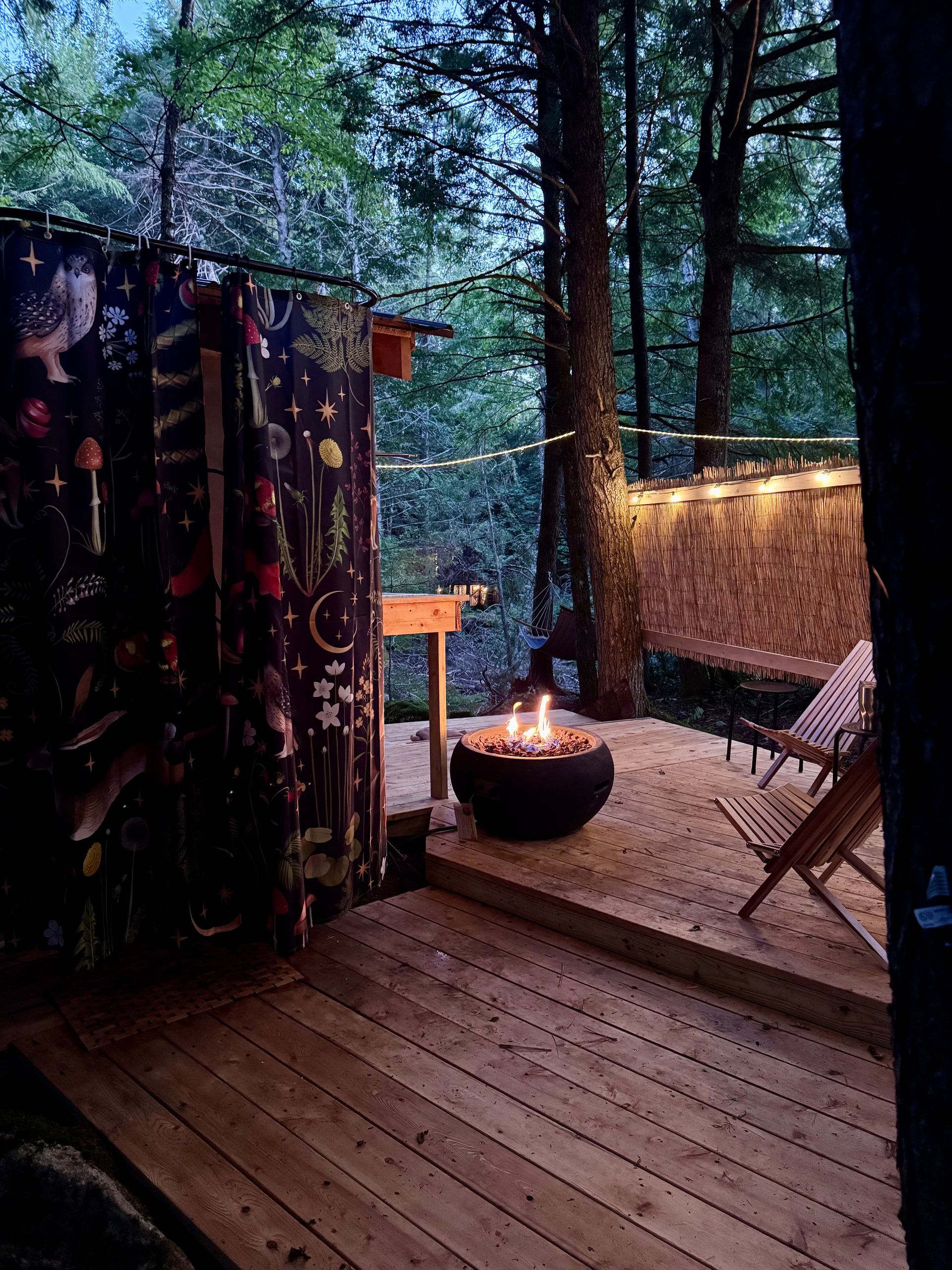 Wooden deck with fire pit, seating, and draped curtain in a forest setting.
