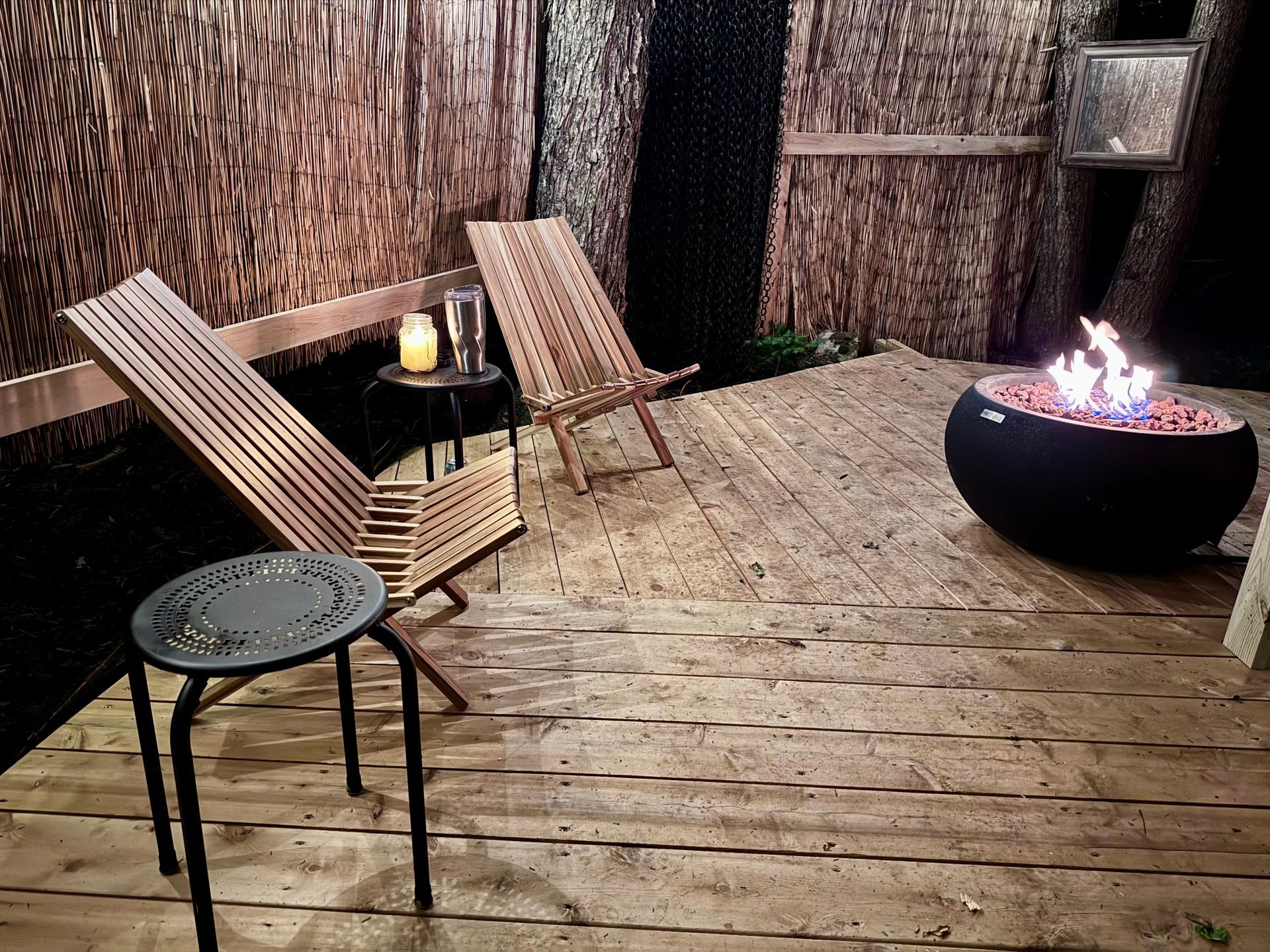 Cozy outdoor seating area with wooden chairs, a small table, fire pit, and reed-like walls.
