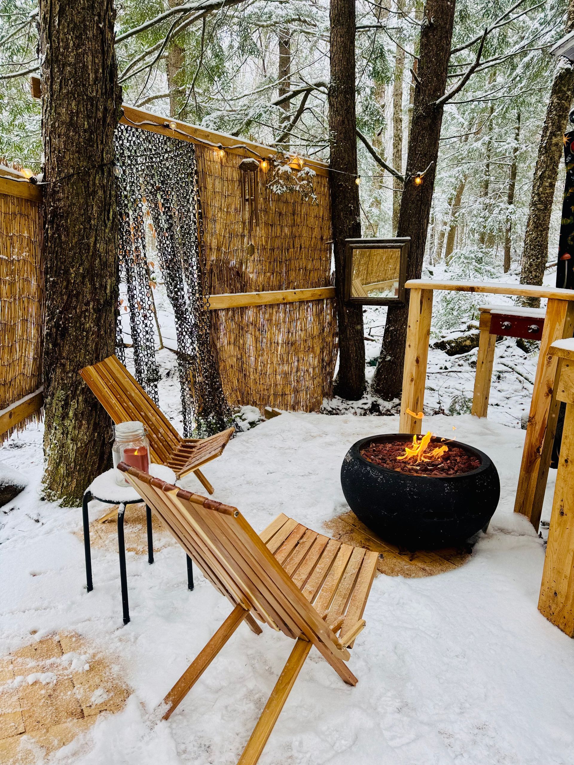 Snowy outdoor seating area with fire pit, wooden chairs, and bamboo fence.