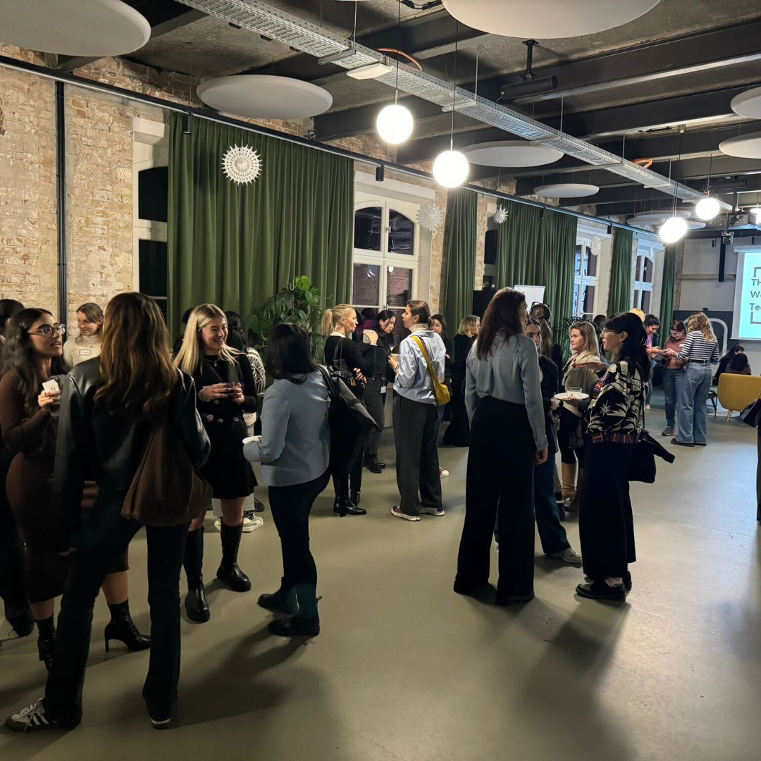 People mingling in a spacious loft venue with brick walls, green curtains, and hanging lights at a social event