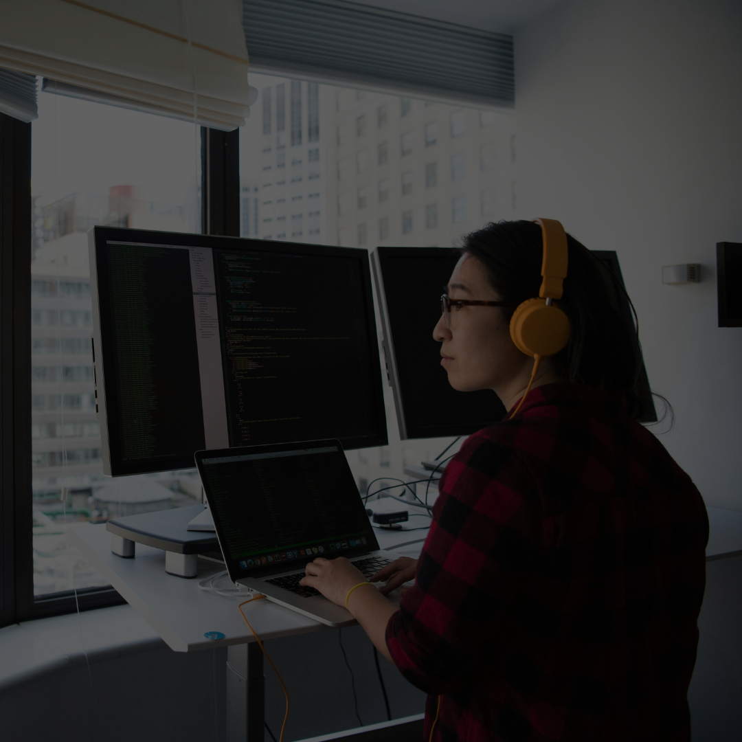 Person in yellow headphones working on a laptop at a desk with dual monitors in a modern office.