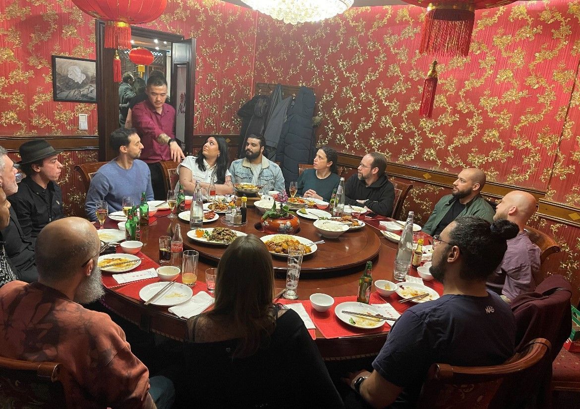 People seated around a crowded dining table in a red wallpapered restaurant, sharing a meal.