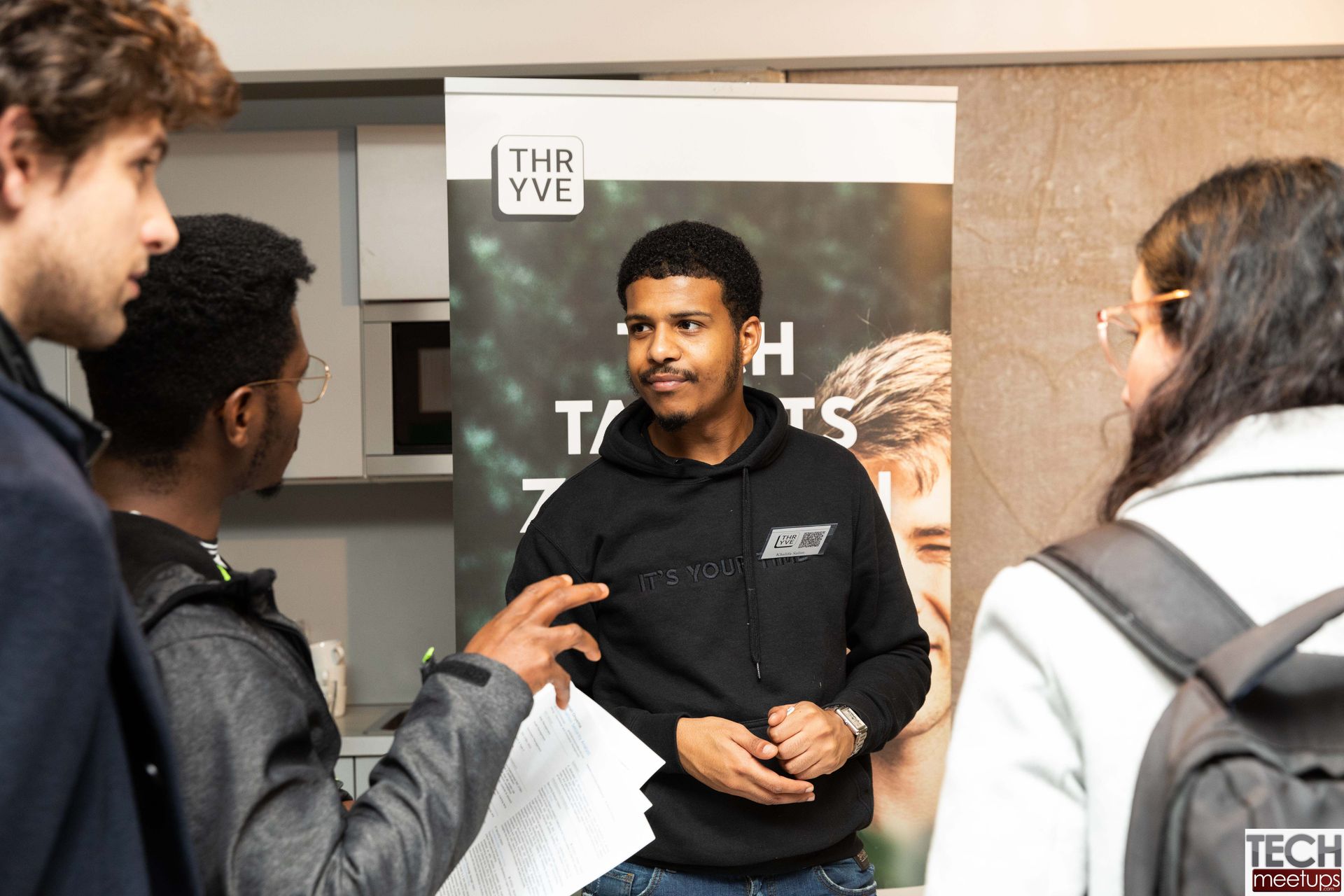 Three people talking at a tech event booth, with one man speaking in front of a banner.