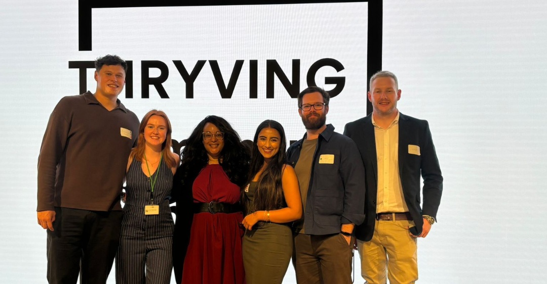 Five people standing on stage in front of a large screen with the word “TRYVING” behind them.