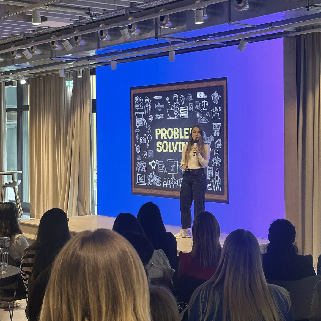 Speaker on stage in a blue-lit room presenting in front of a chalkboard-style “Problem Solver” backdrop.