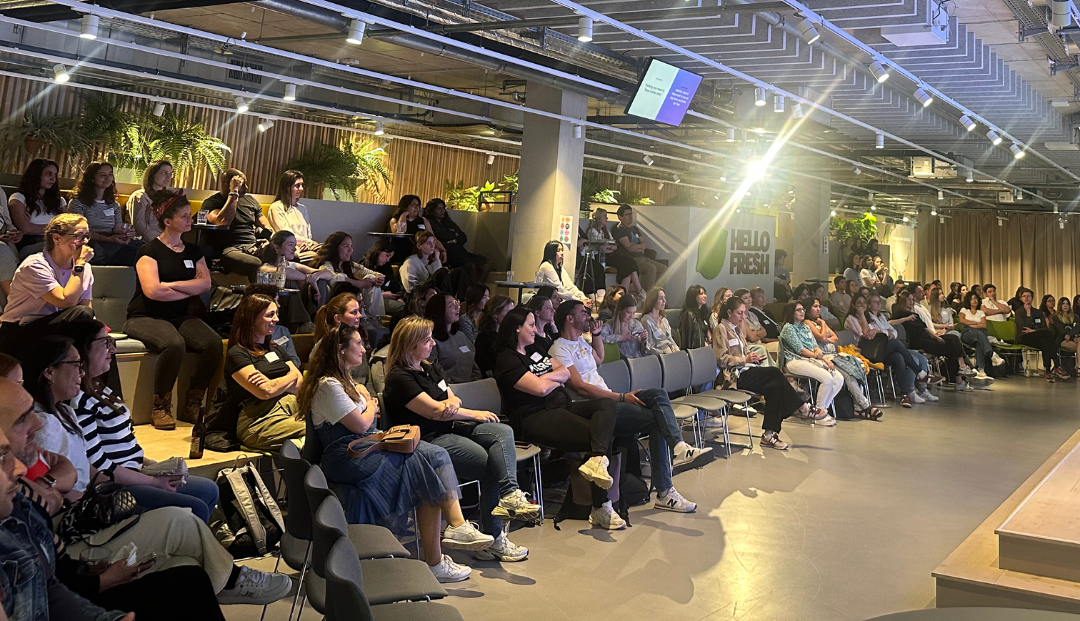 Crowd seated in an industrial-style venue, watching a bright presentation on a large screen.