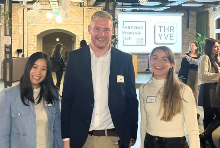 Three people smiling at an indoor event, with Thrive and Women Tech signs in the background.