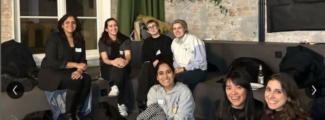 Six people smiling on a couch in a dim indoor living room, with a laptop in front.
