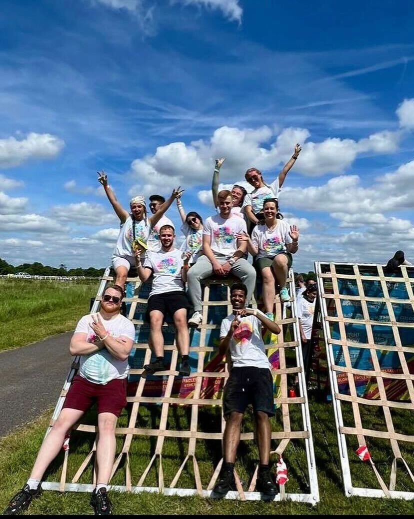 Group of people posing on a wooden climbing frame outdoors under a bright blue sky.