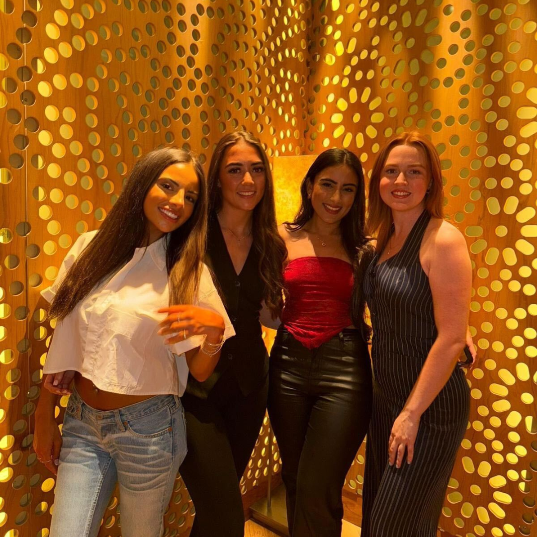 Four friends posing in front of a gold polka-dot backdrop, wearing festive outfits and smiling.
