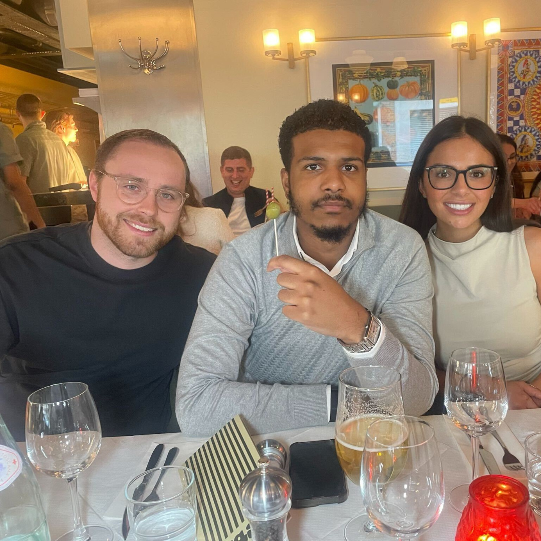 Three friends smiling at a restaurant table, with glasses and a red candle in the foreground.