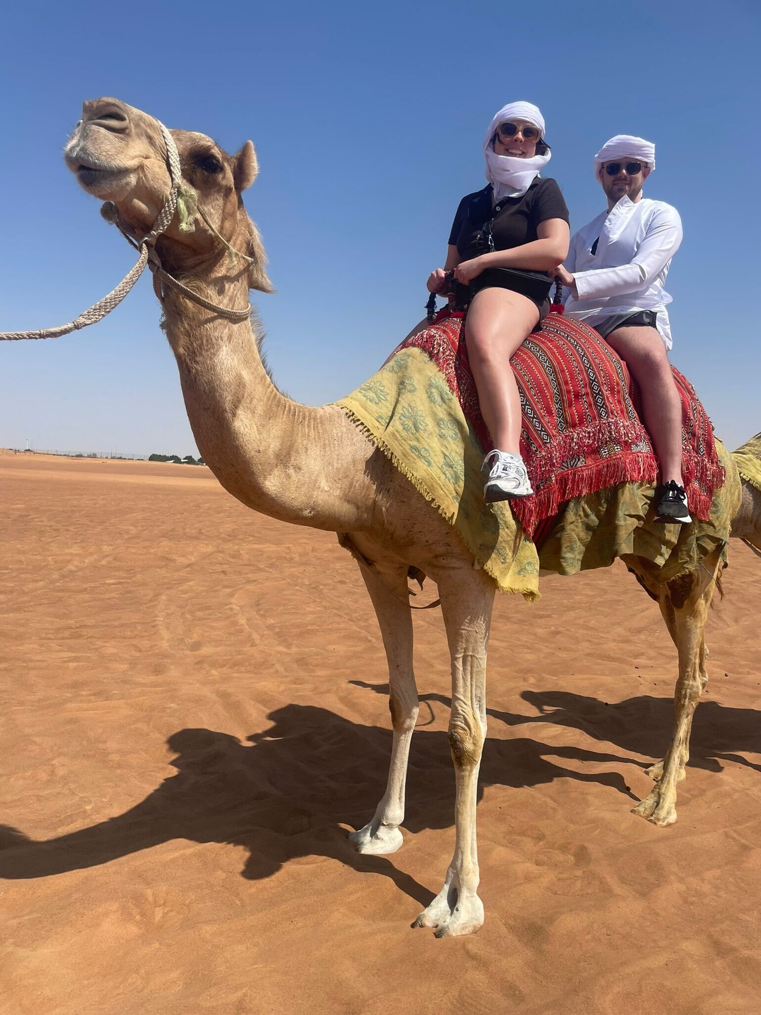 Two masked riders on a camel in a desert, with red saddle blankets and a bright blue sky.