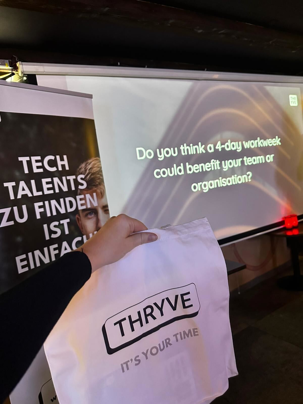 Person holding a white THRYVE bag in front of event banners with German and English text
