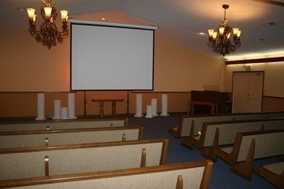 Empty church hall with rows of pews facing a projection screen and altar area under chandeliers.