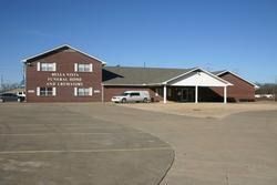 Red brick school building with a white entrance, parked cars, and a large empty parking lot under a clear sky