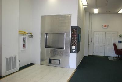 Lobby with a stainless steel wall fixture, white walls, and a hallway leading to a door with red chairs.