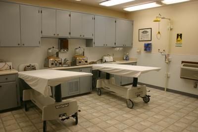 Medical exam room with two covered exam tables, cabinets, and a sink on a tiled floor.