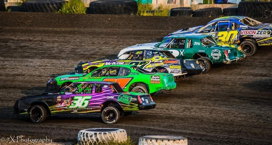 Colorful stock cars racing closely through a dirt track turn, led by a purple and green car with number 75.