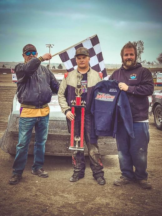 Three people outdoors, one holding a checkered flag and one holding a racing trophy and jacket.