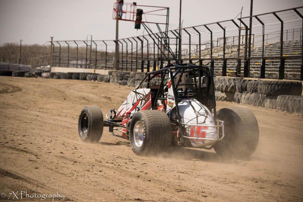 Open-wheel race buggy kicking up dust on a dirt track beside safety fencing
