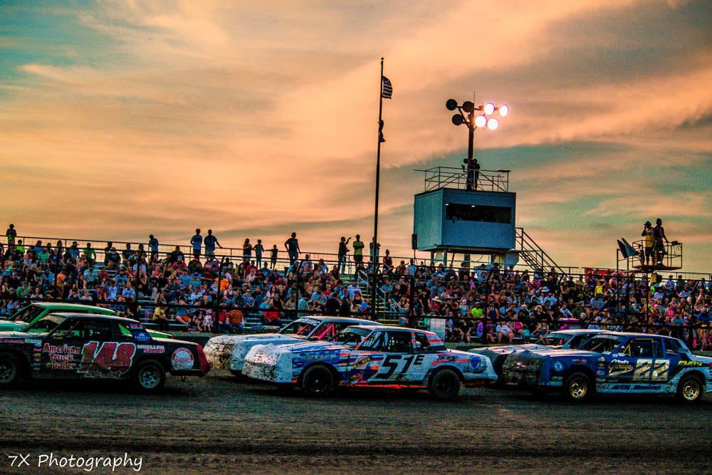 Stock cars lined up on a dirt track at sunset, with a crowded grandstand and scoreboard in the background