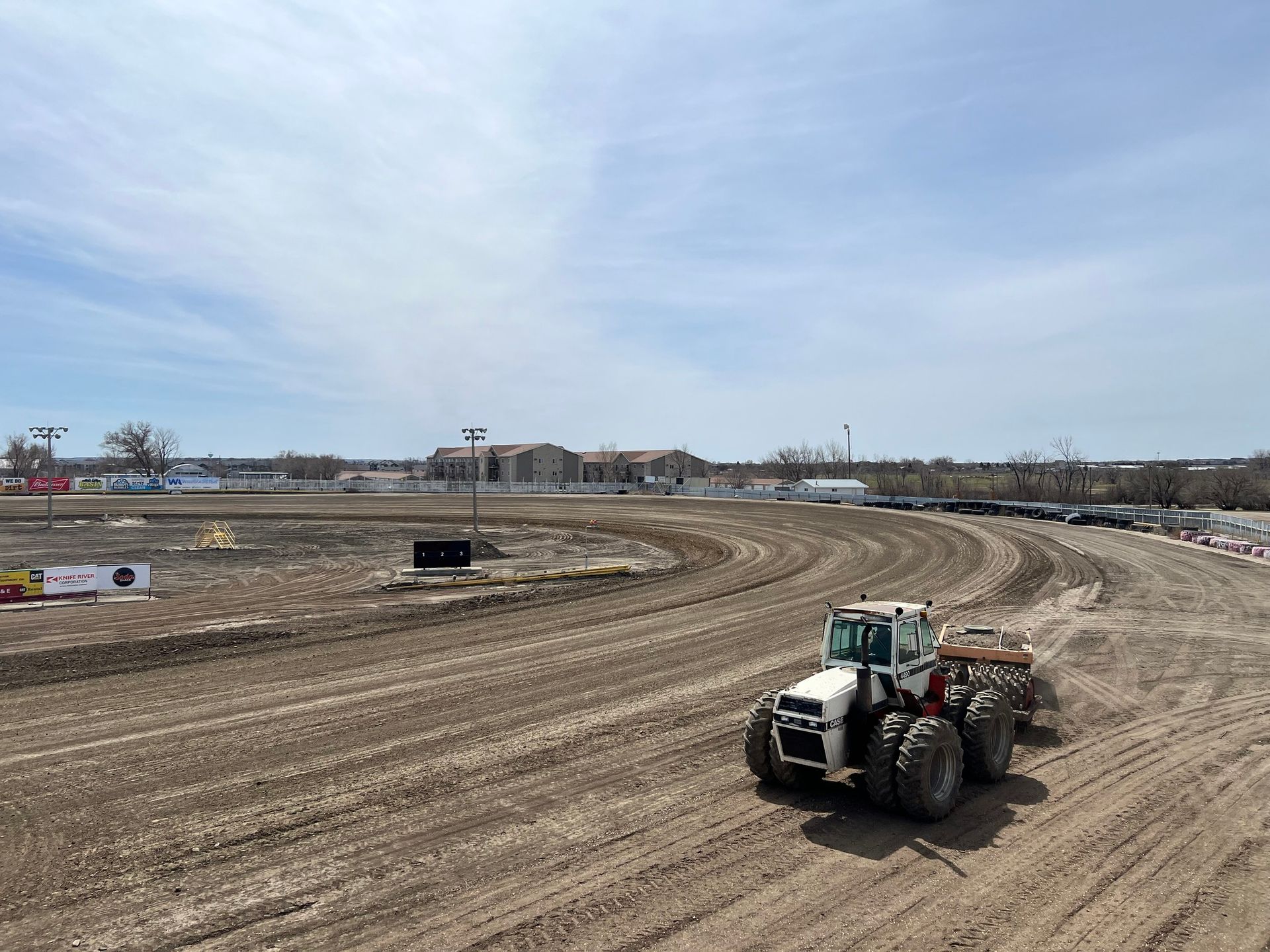 Tractor grading a dirt racetrack under a cloudy sky, with a curved track and empty stands in the distance