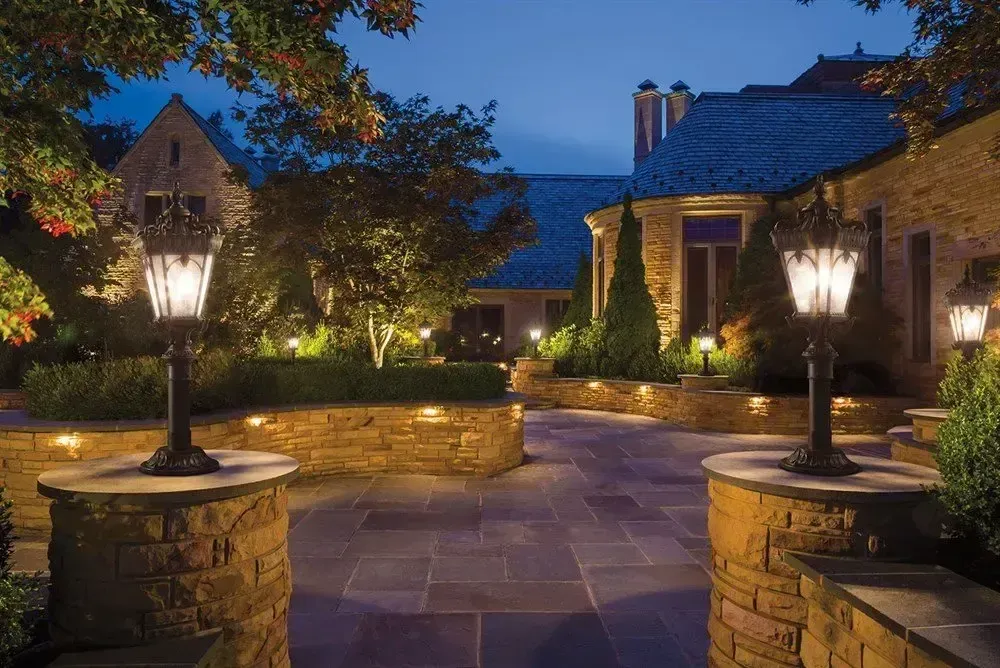 Lit courtyard at dusk with stone walls, steps, and lampposts. House in background.