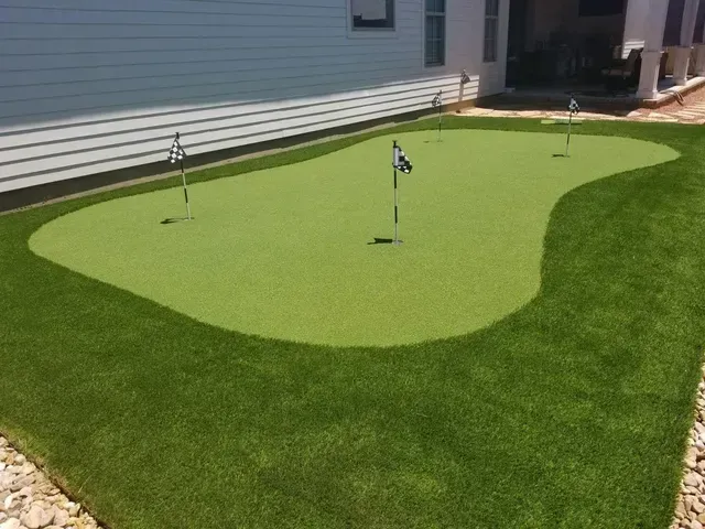 Artificial putting green in a backyard with several flags. Green grass and border with rocks.