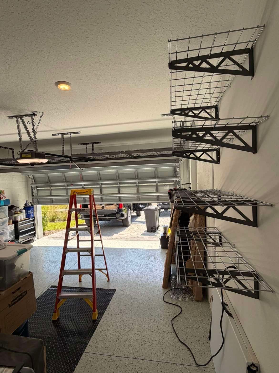 Garage interior with shelves on the right, ladders, tools, and an open garage door letting in daylight