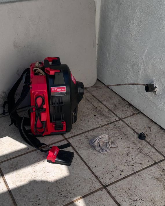 Red and black backpack-style machine on tiled floor against wall, with a small plug and cord nearby.
