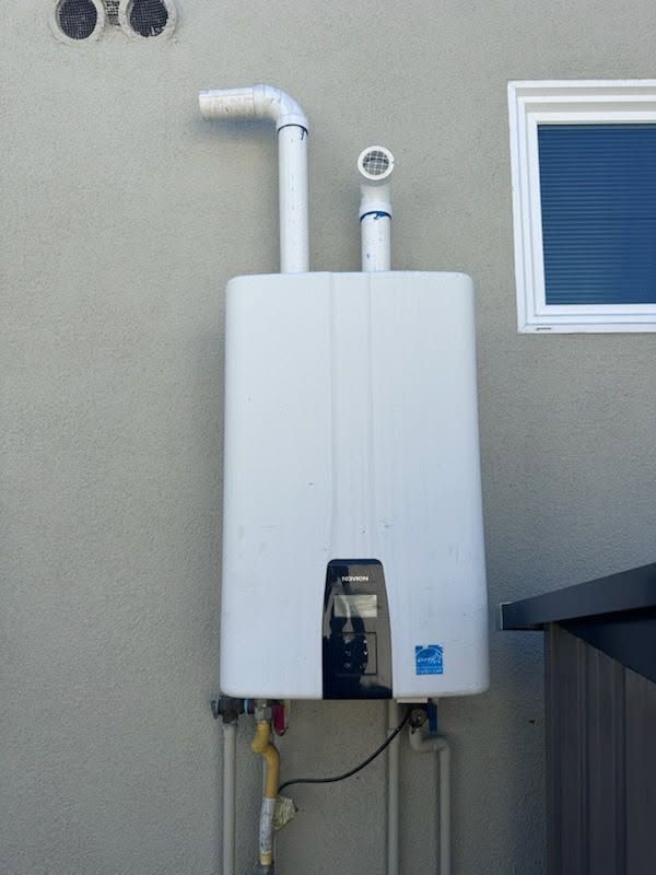White wall-mounted tankless water heater with exposed pipes on an exterior beige wall.