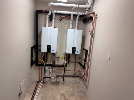 Utility room with two wall-mounted water heaters, exposed pipes, and beige walls and floor.
