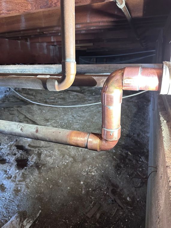 Rusty copper pipes under a floor, with elbows and joints against a dark, dirty surface
