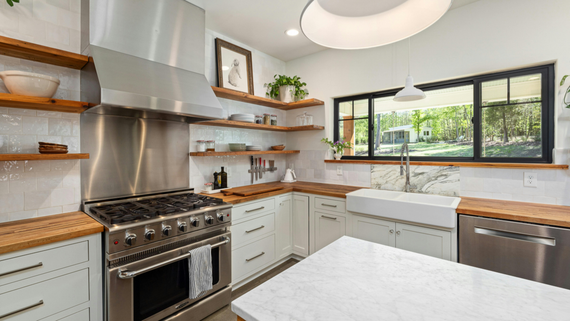 Modern kitchen with stainless steel stove, white cabinets, wooden shelves, and large window over the sink