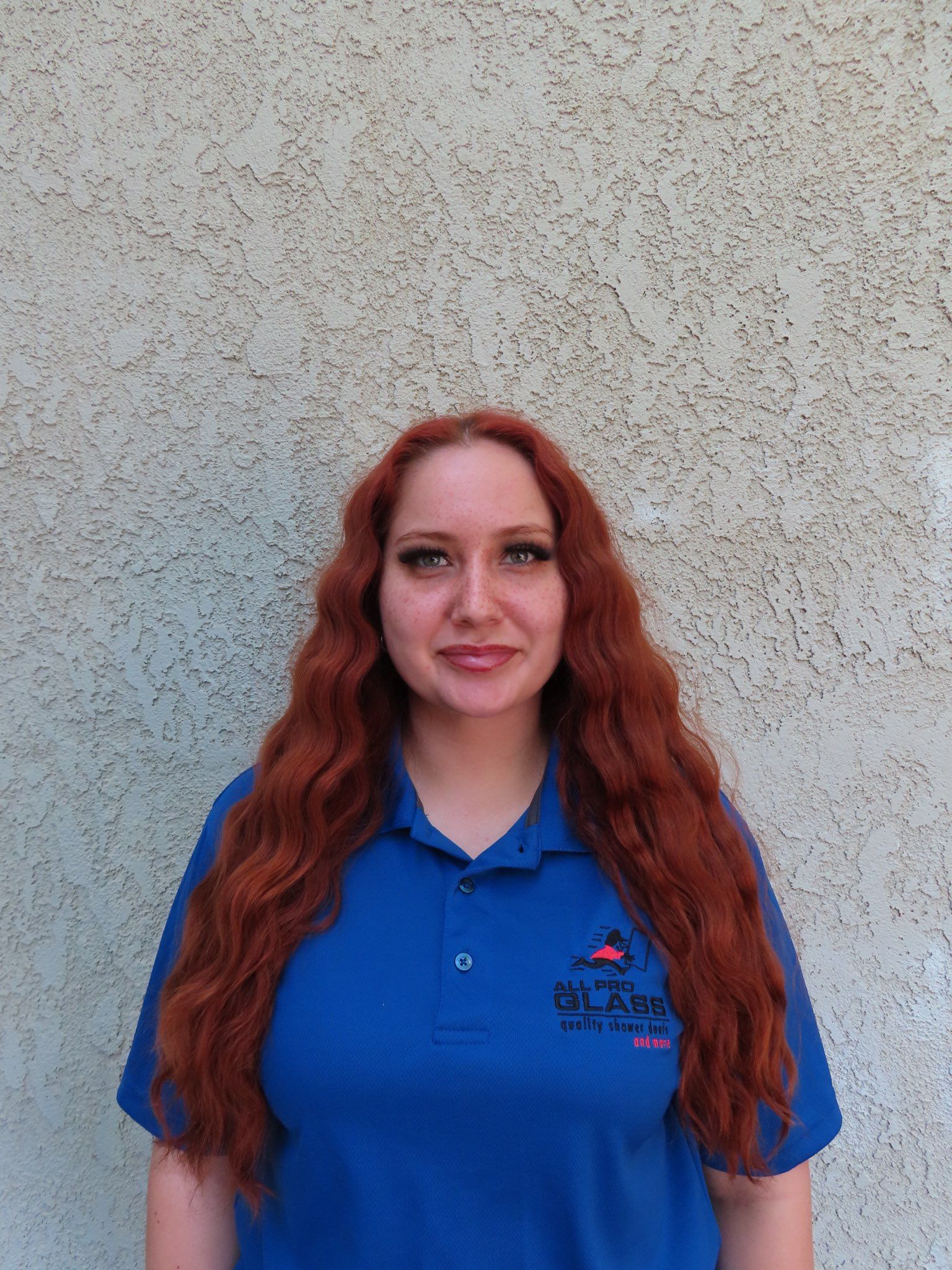 Woman with long red hair wearing a blue polo, smiling against a speckled wall.