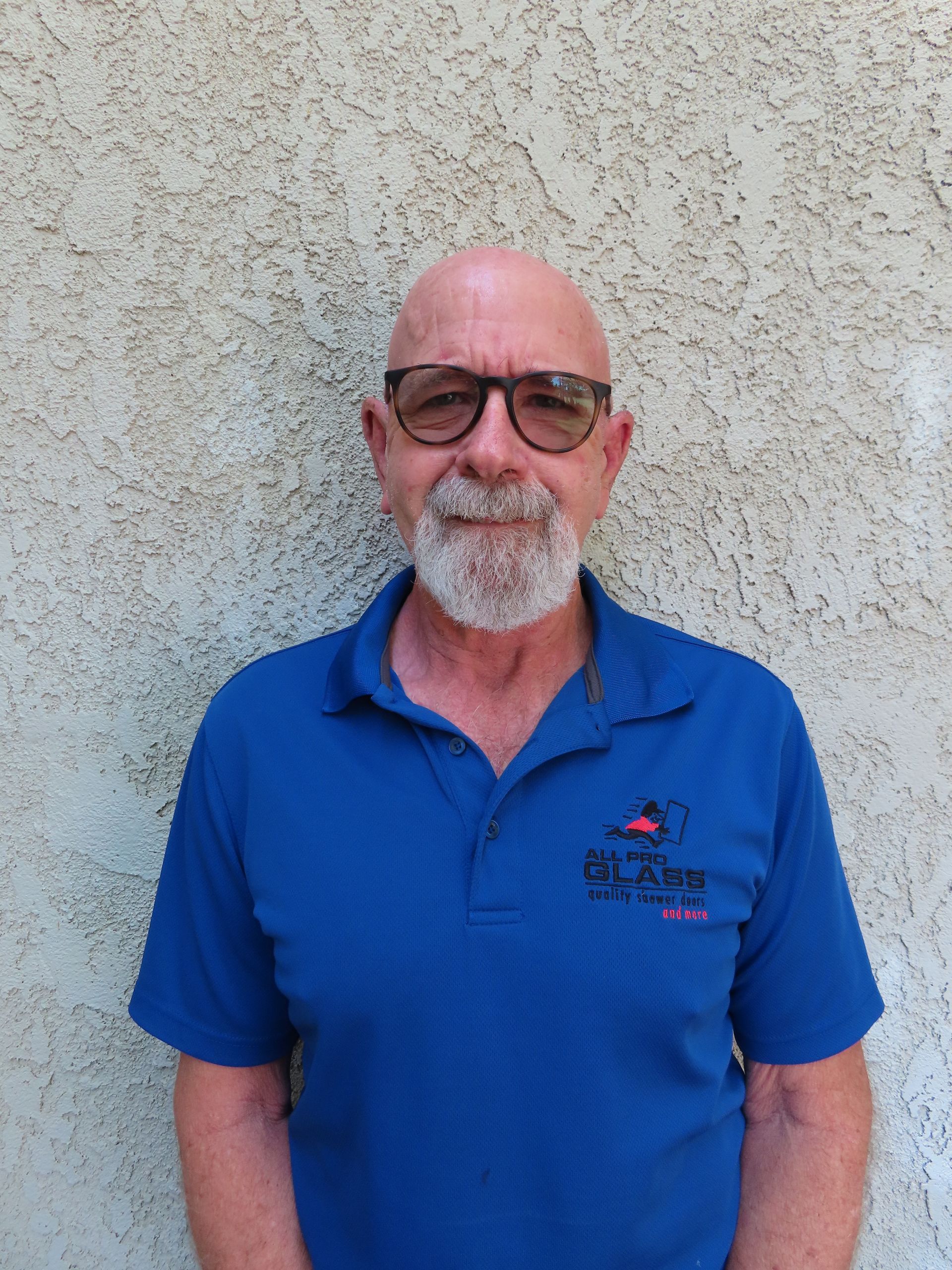 Man standing against a textured wall, wearing a blue polo shirt and glasses, with a gray beard.