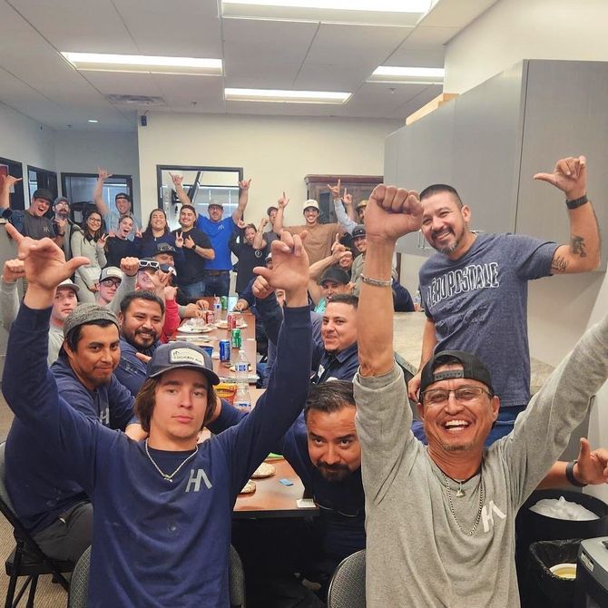 Group of people cheering in a room, arms raised. Many wear work attire, and some have caps.