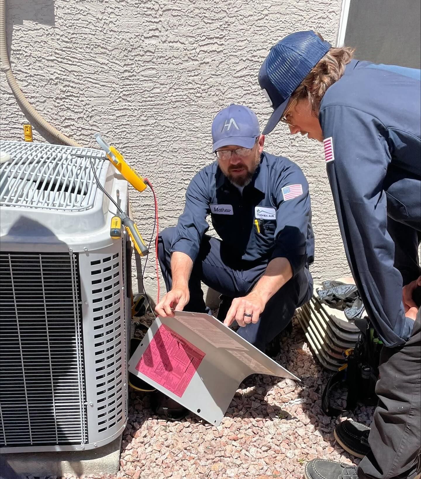 Two HVAC technicians examining an air conditioner unit outside. One points at a component.