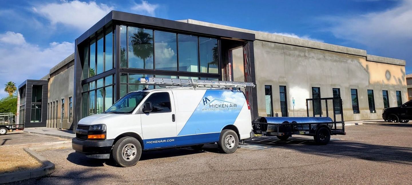 White work van with logo parked in front of a modern building with large glass windows. Trailer attached. Blue sky.