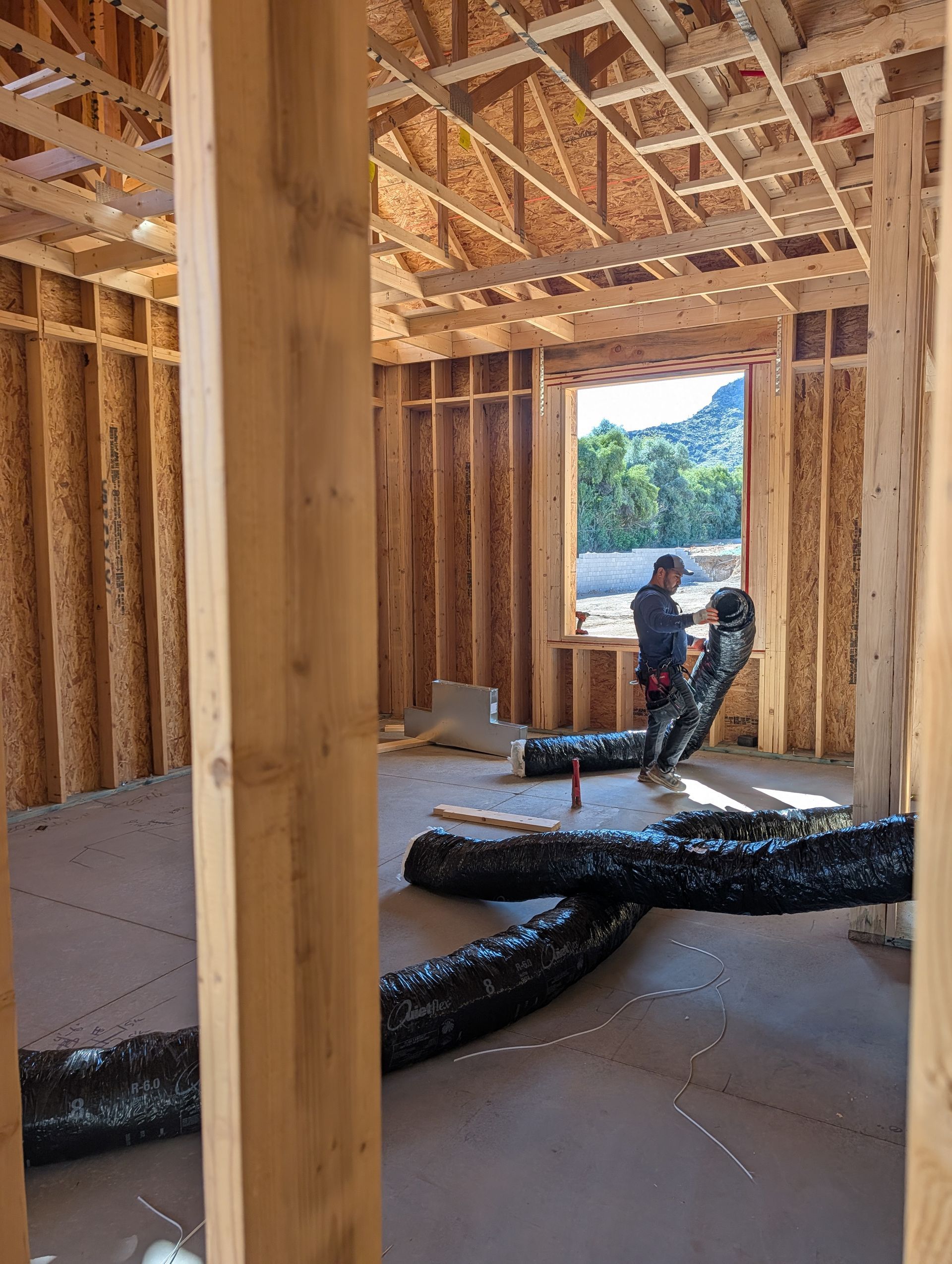 Wood-frame house under construction with two workers, exposed insulation, and mountain view through an open doorway