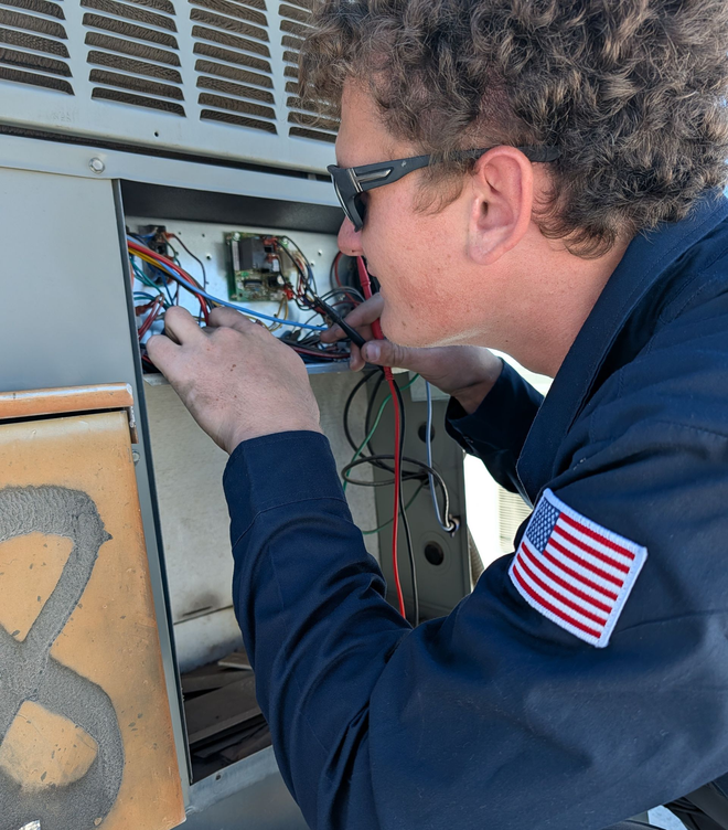 Two technicians working on an AC unit outside. One standing, one kneeling, both in navy uniforms.