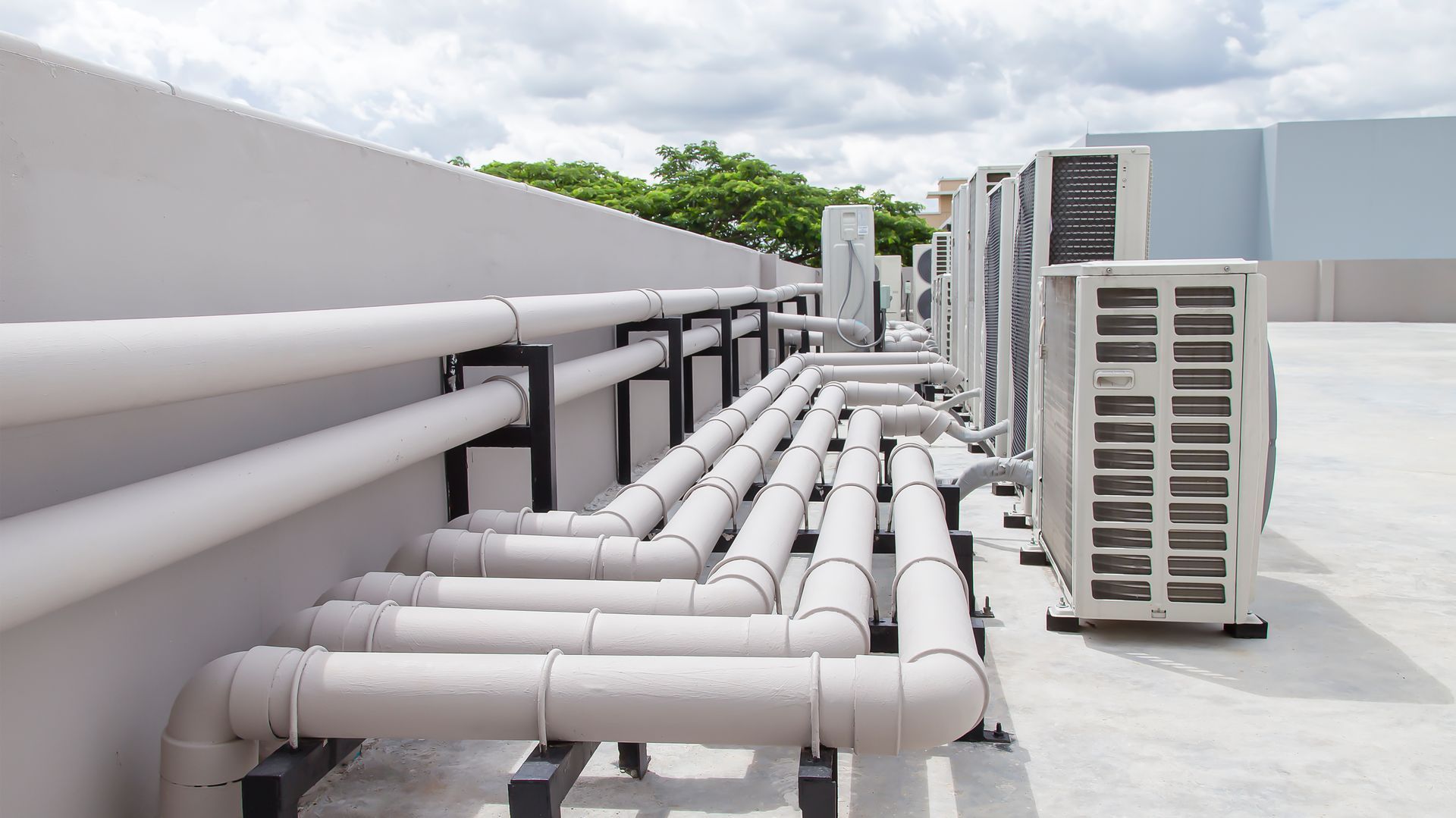 Air conditioning units and white pipes on a rooftop against a cloudy sky.