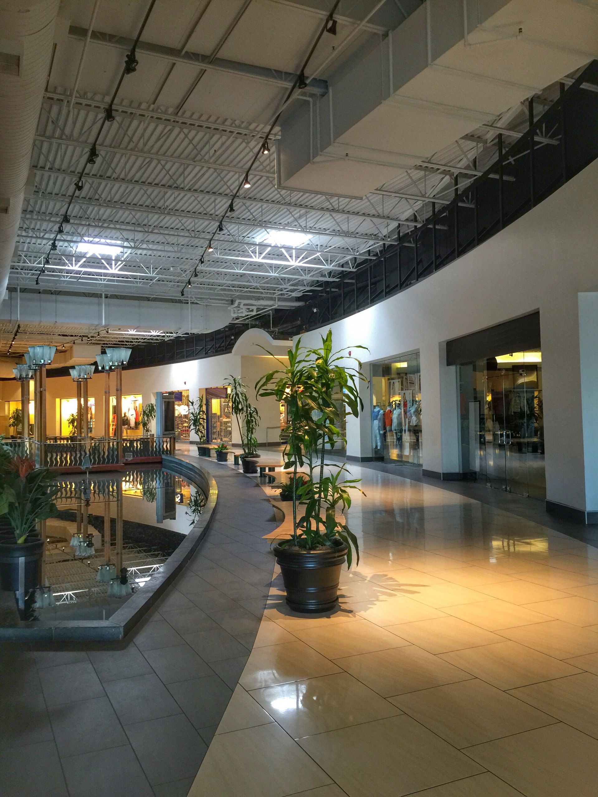 Interior of a shopping mall. Long hallway with stores, potted plants, and water feature.