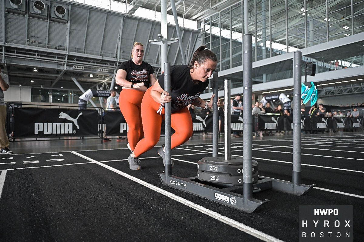 Athletes pushing a sled obstacle on an indoor track, one in red leggings and black shirt.