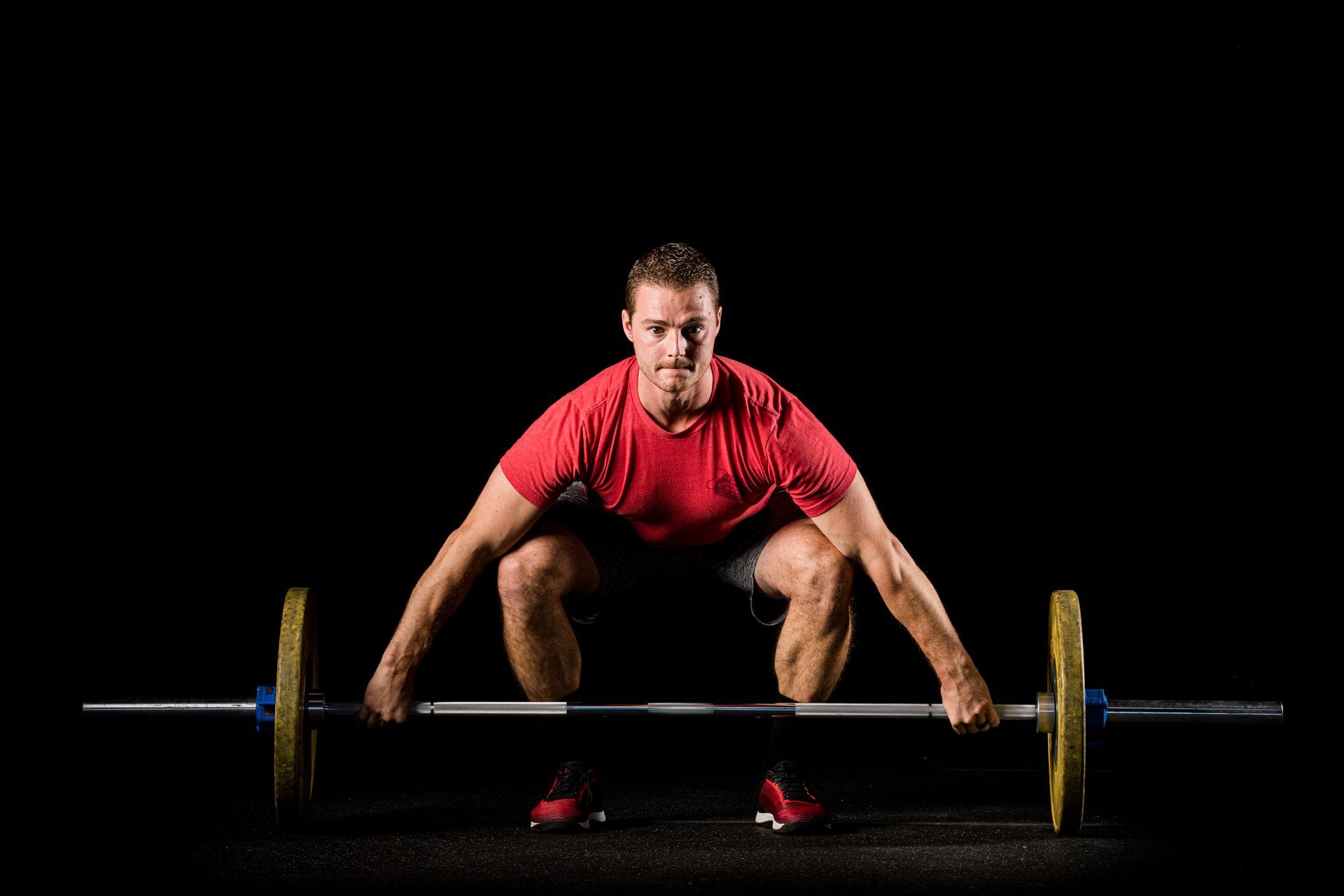 Athlete in red shirt crouches to lift a barbell on a dark stage