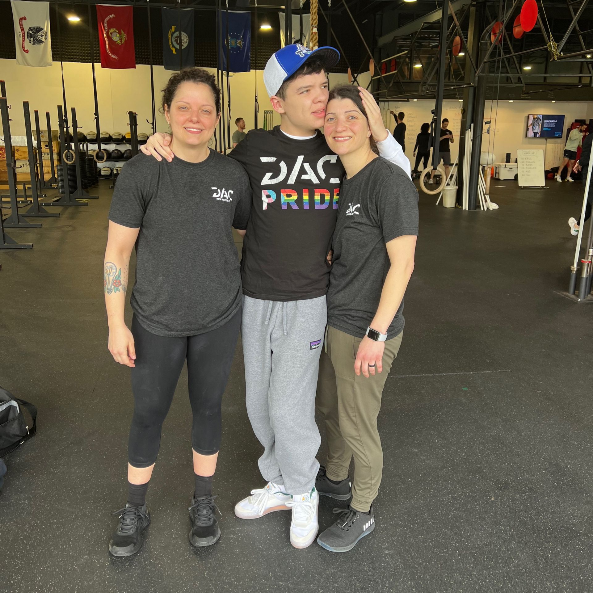 Three people posing together in a gym, smiling and standing close, with flags and exercise equipment behind them