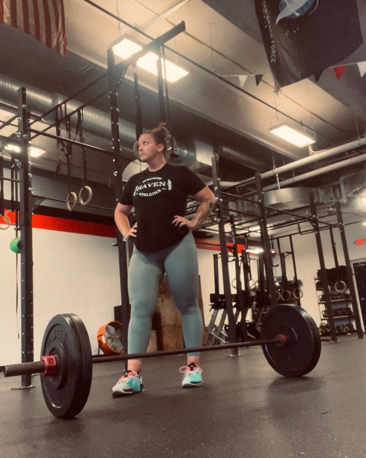 Womanat Strength Haven Athletics gym, arms on hips, stands over barbell. She's wearing blue leggings and a black shirt.