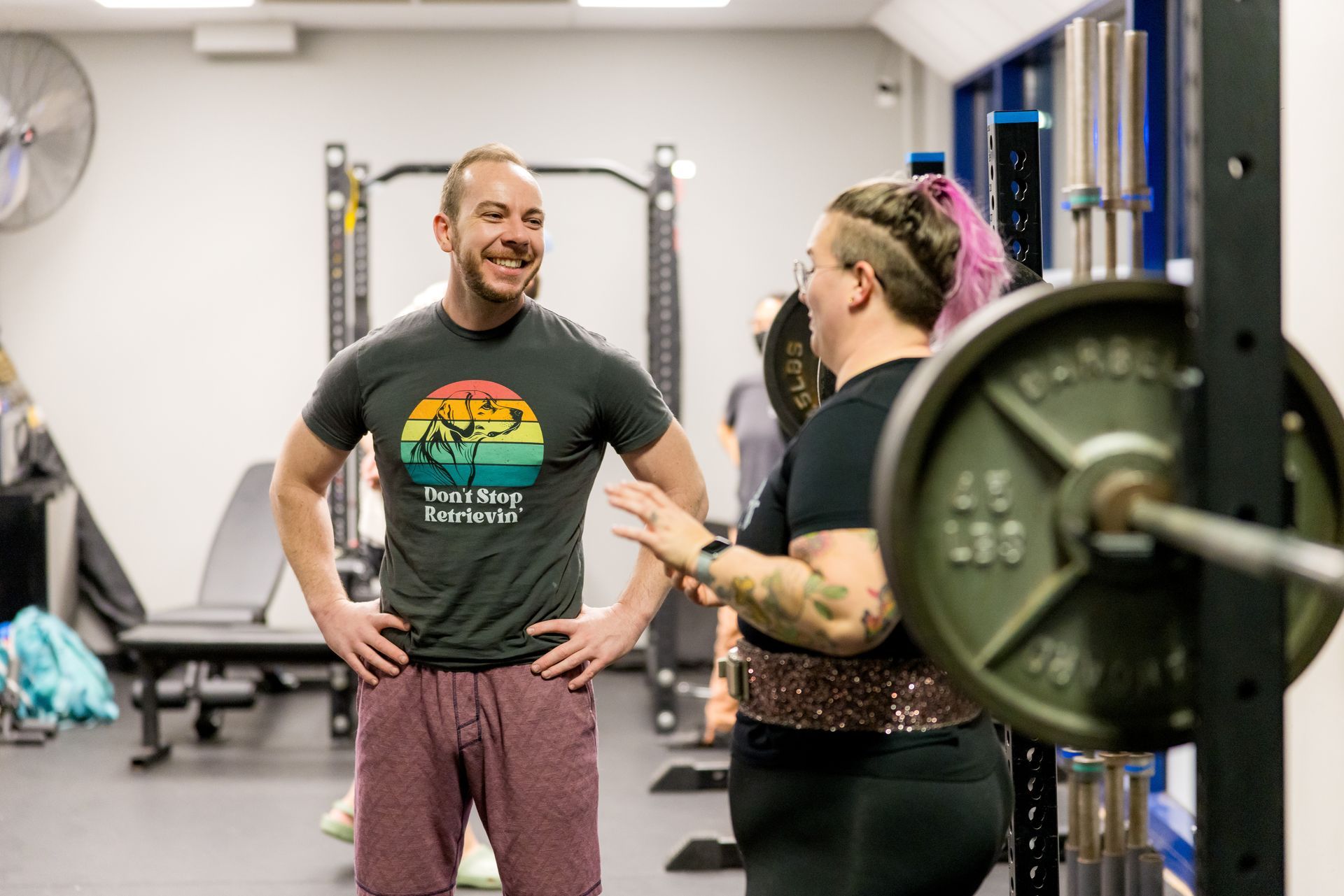 Man and woman lifting partners at a gym, smiling during a weightlifting session. Woman has arm tattoos and weight on the bar.