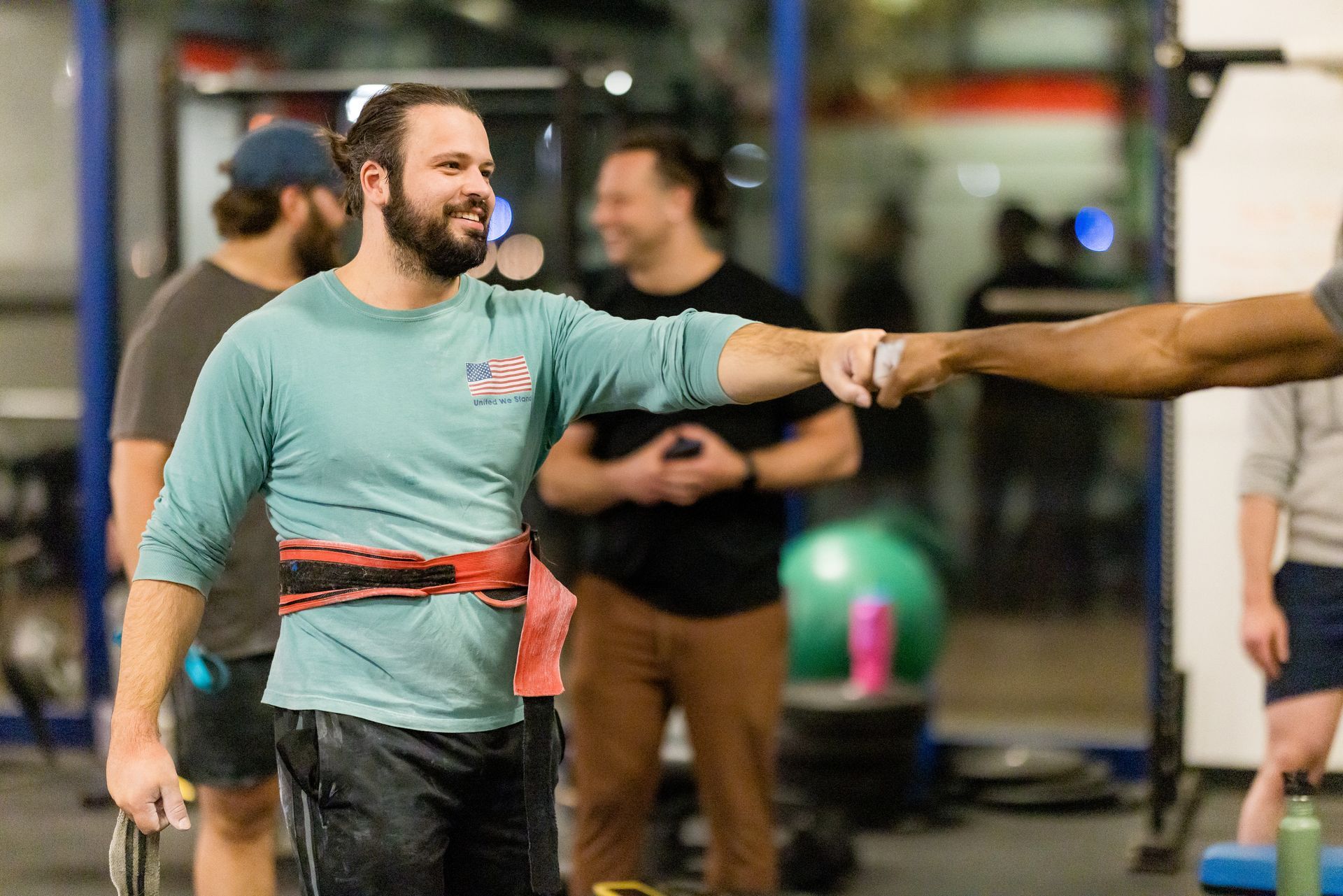 Coach in blue shirt and weight belt greets member at Strength Haven, with gym community active in the background